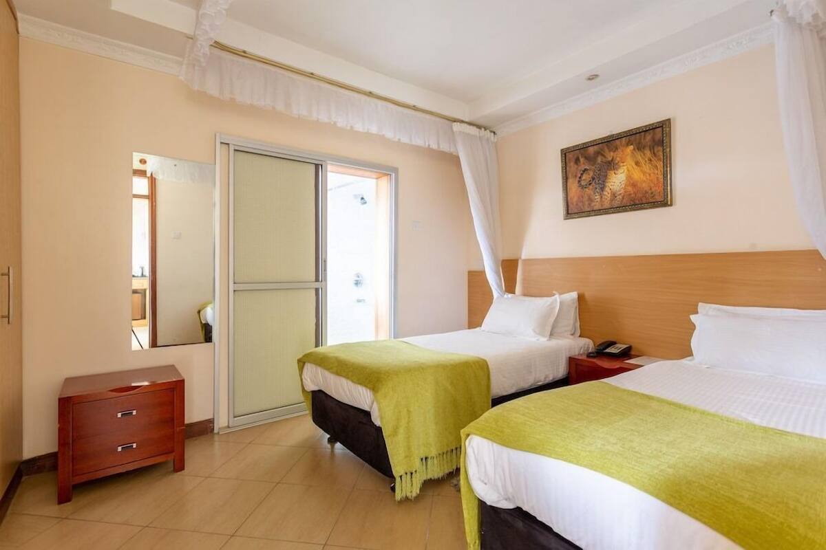 A hotel room with two beds, a wooden headboard, a bedside table with a phone, a small wooden cabinet, and a sliding glass door.
