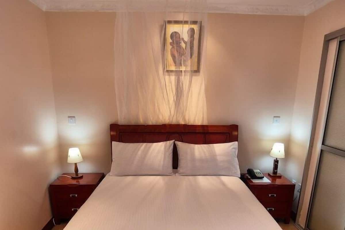 A bedroom with a bed, two bedside tables, a nightstand, a green rug, a hanging light fixture, and a framed picture on the wall.