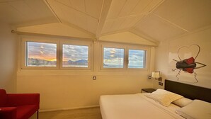 A hotel room with a bed, a red chair, and three windows with a cityscape view. - Hotel Punta Monpás (San Sebastián)