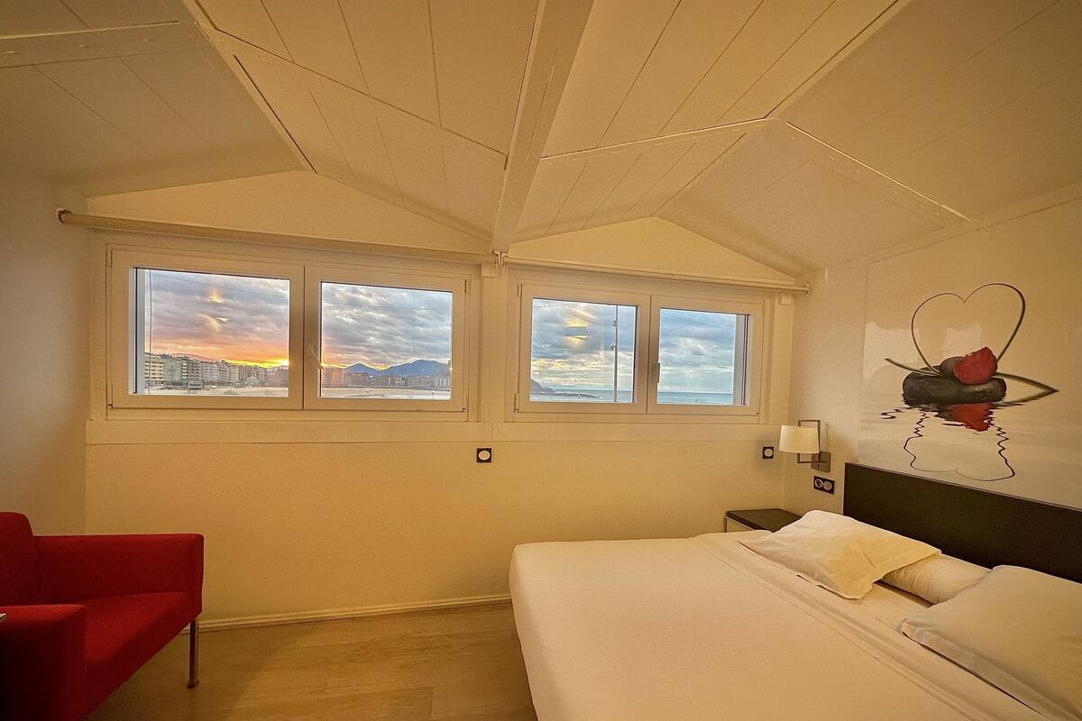A hotel room with a bed, a red chair, and three windows with a cityscape view.