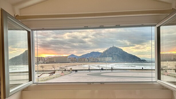 A room with a large window overlooking a beach, a red chair, and a white table. - Hotel Punta Monpás (San Sebastián)