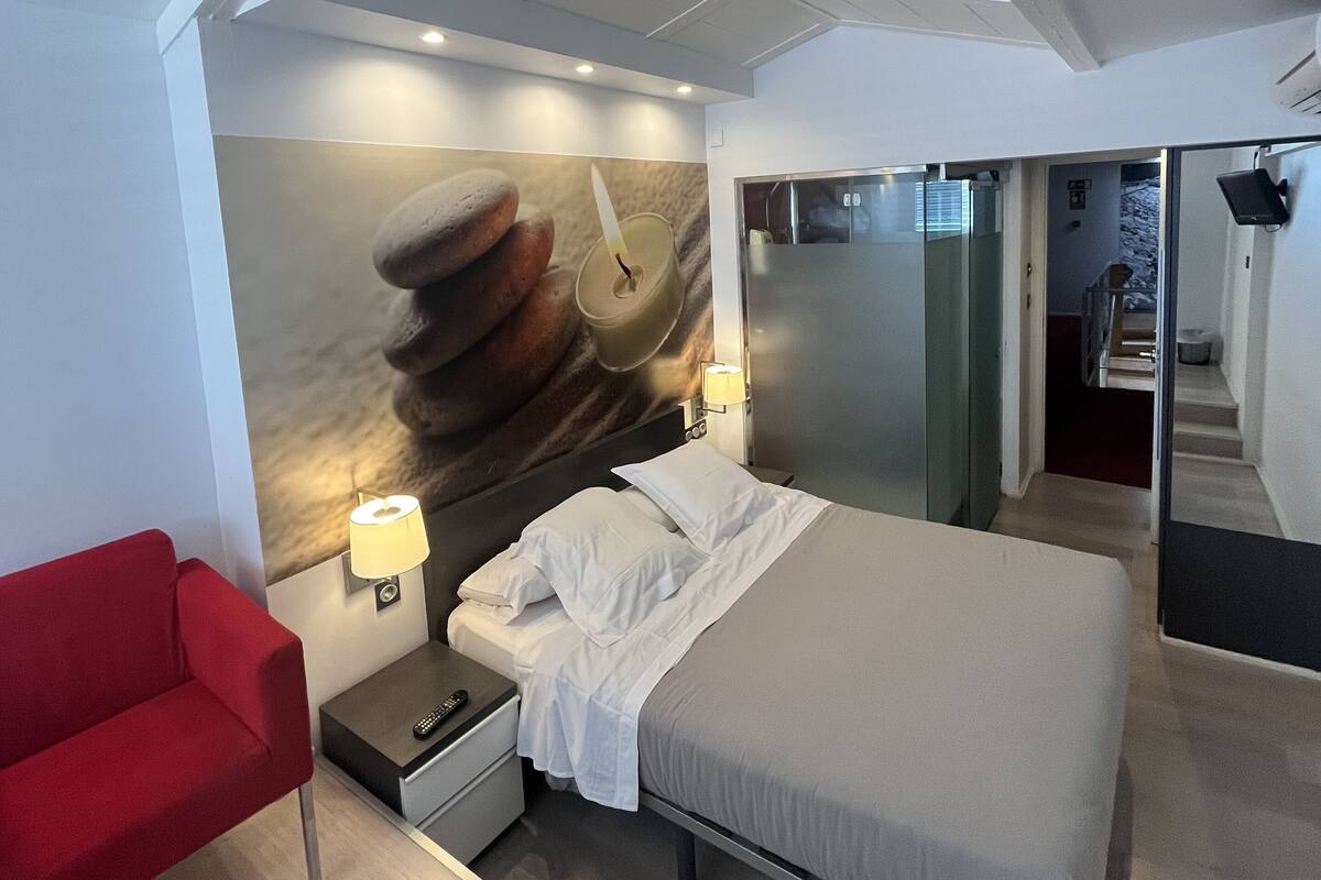 A modern hotel room with a bed, a red chair, a nightstand, a wall-mounted lamp, and a large abstract painting.