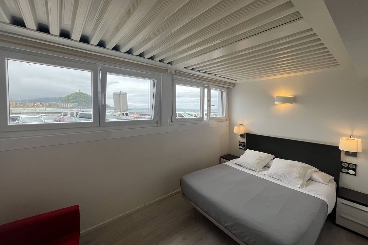 A hotel room with a bed, bedside lamps, a nightstand, a red chair, and a view of the city through large windows.