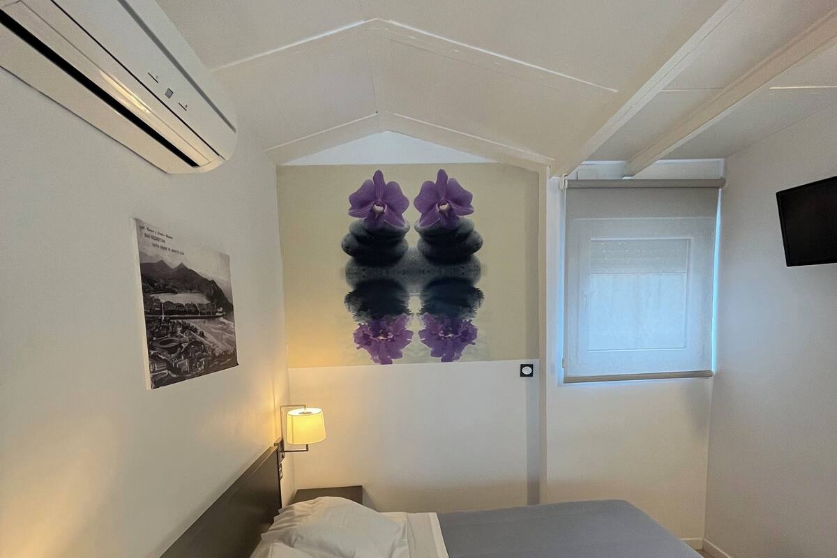 A hotel room with a bed, a nightstand with a lamp, a wall-mounted air conditioner, a framed picture, a window with a view, and a wall decoration of purple flowers.
