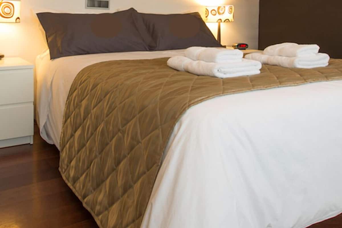 A neatly made bed with a brown quilt and white linens, two white towels on the bed, and a bedside table with a lamp and a clock.
