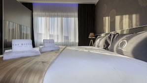 A hotel room with a large bed, two towels with "The Queen Luxury Living" branding, a nightstand, and a view of the city through sheer curtains. - The Queen Luxury Apartments - Villa Giada (Hesperange)