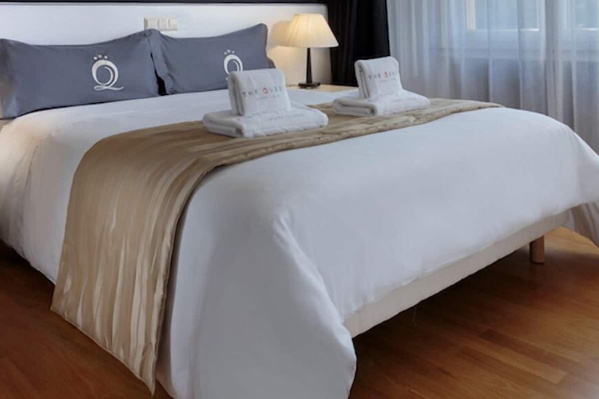 A neatly made bed with white linens, a beige throw blanket, and two folded towels with the hotel logo.