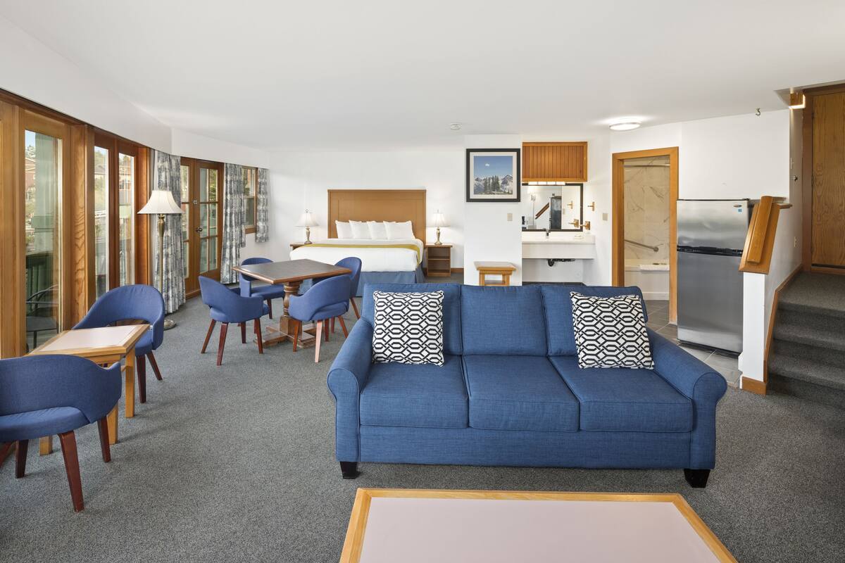A modern hotel room with a blue sofa, dining table, chairs, a bed, a kitchenette, and large windows offering a view.