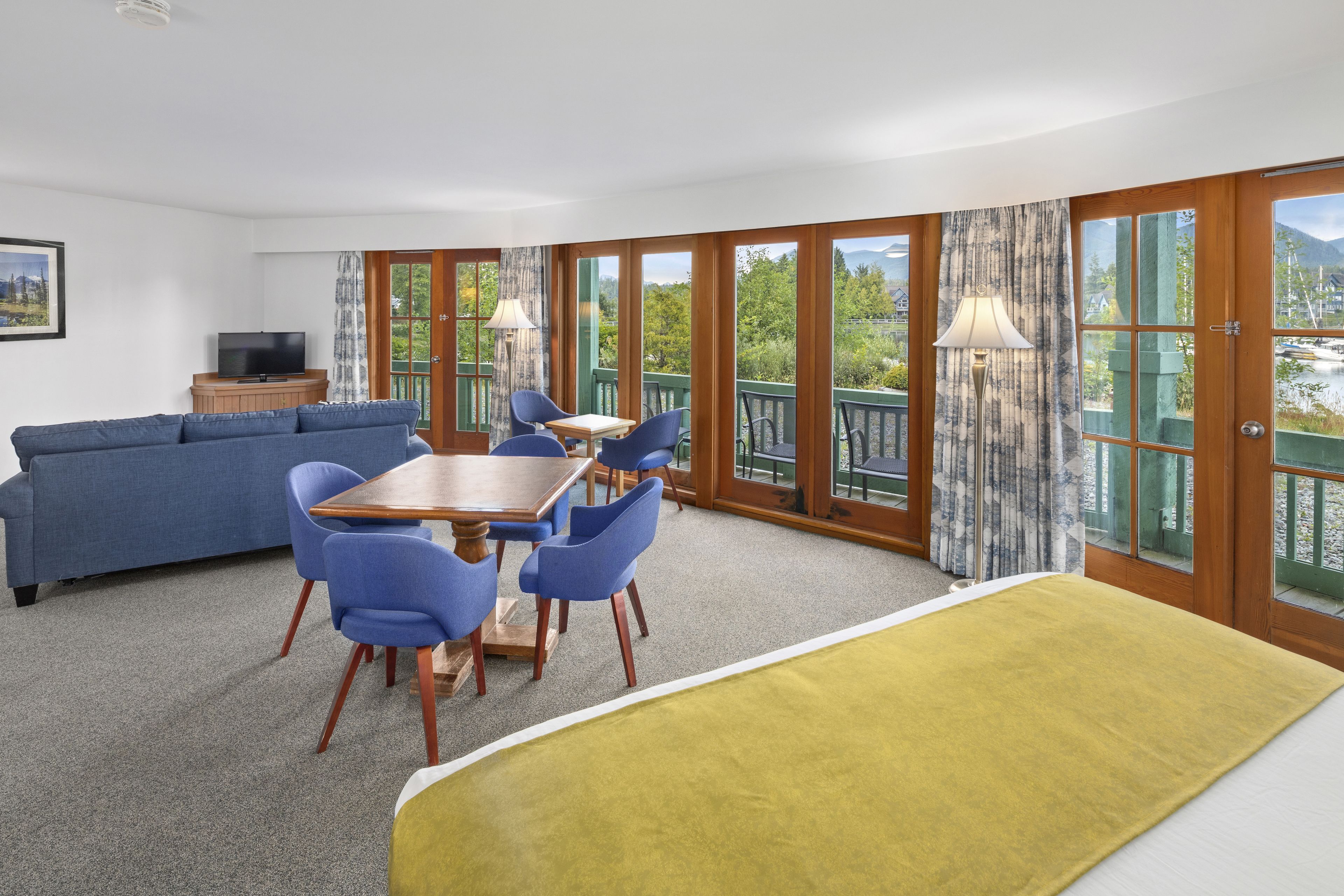 A room with a sofa, dining table, chairs, a TV, and large windows with a view of greenery and a body of water.