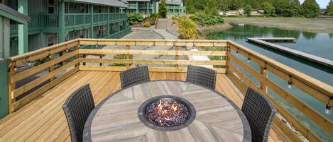 A wooden deck with a circular table and chairs, a fire pit, and a view of a lake and buildings in the background.