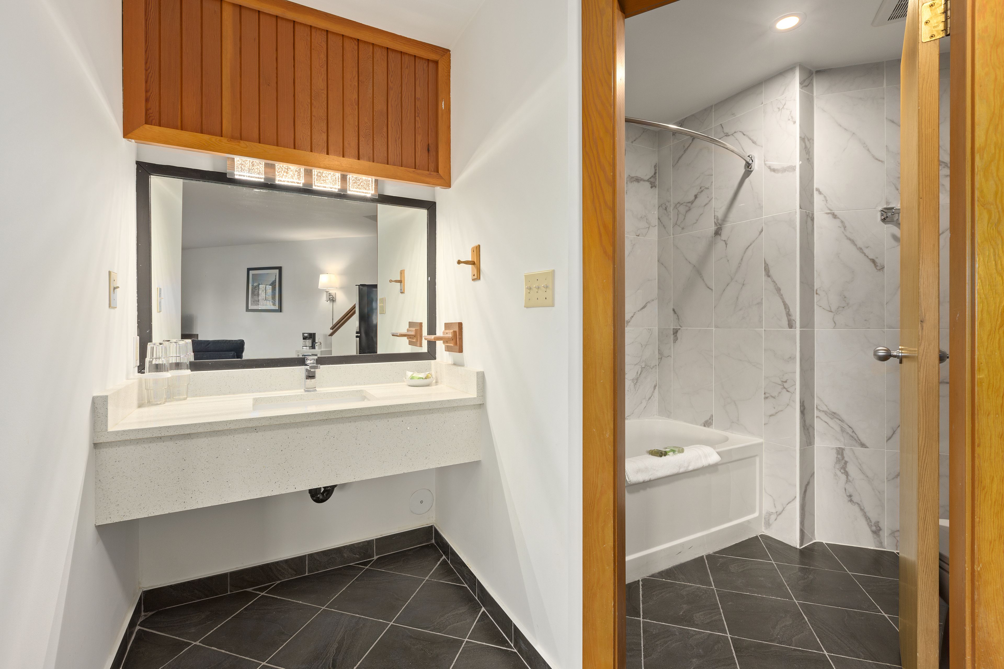 A bathroom with a white vanity, a mirror, a wooden cabinet, a bathtub, and a shower with marble tiles.