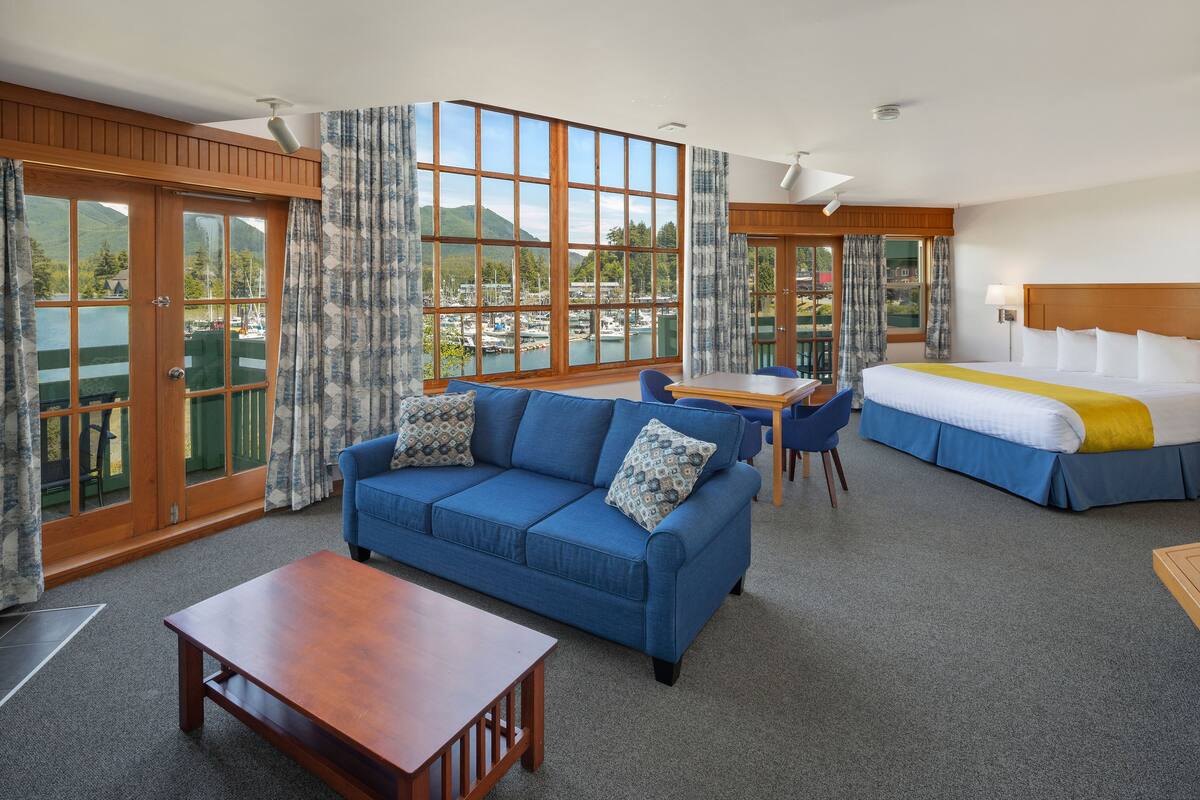 A hotel room with a blue sofa, a wooden coffee table, a bed with a blue and white bedspread, and large windows offering a view of a marina and mountains.