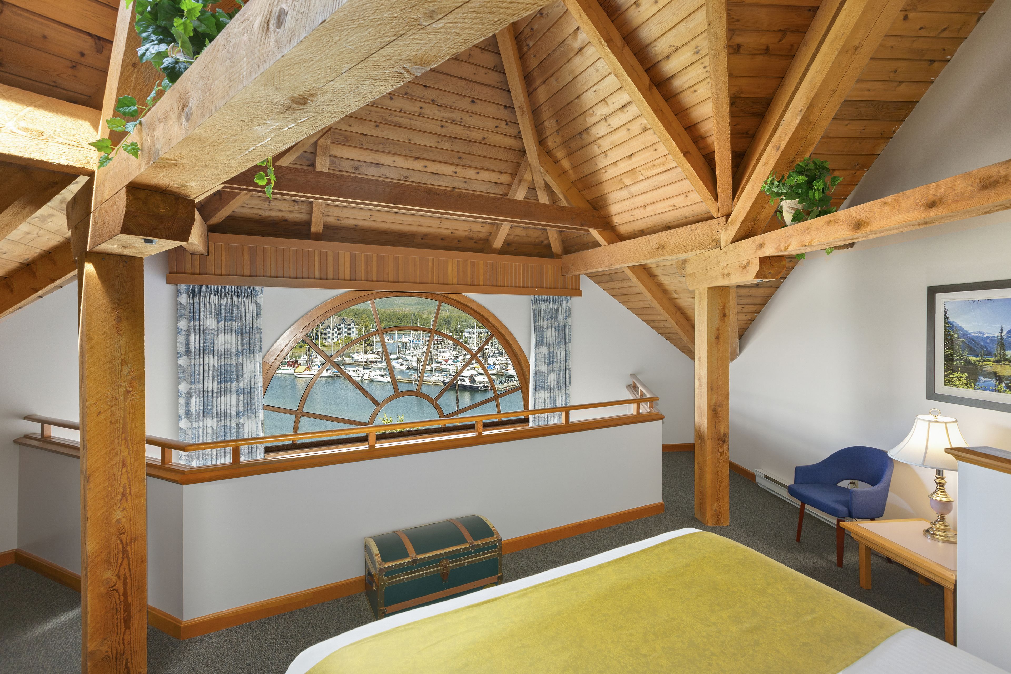 A room with a wooden ceiling, a window with a view of a marina, a bed, a chair, and a desk with a lamp.