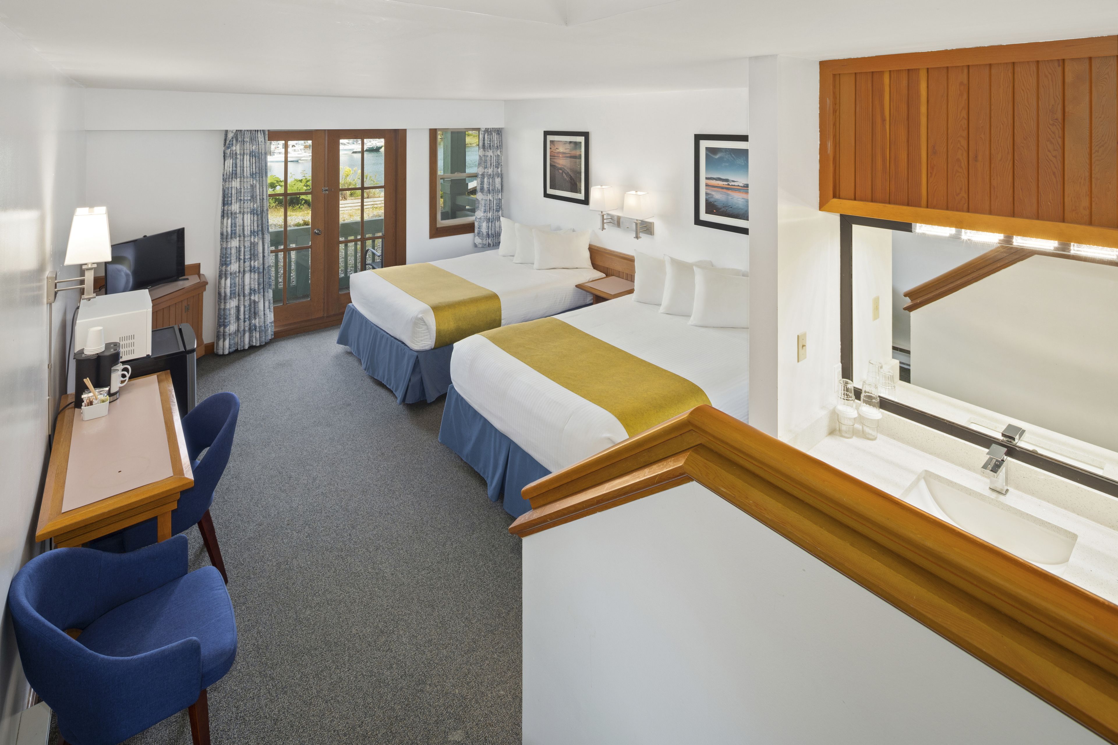 A hotel room with two beds, a desk with a computer, a chair, a large window with curtains, and a wooden ceiling.