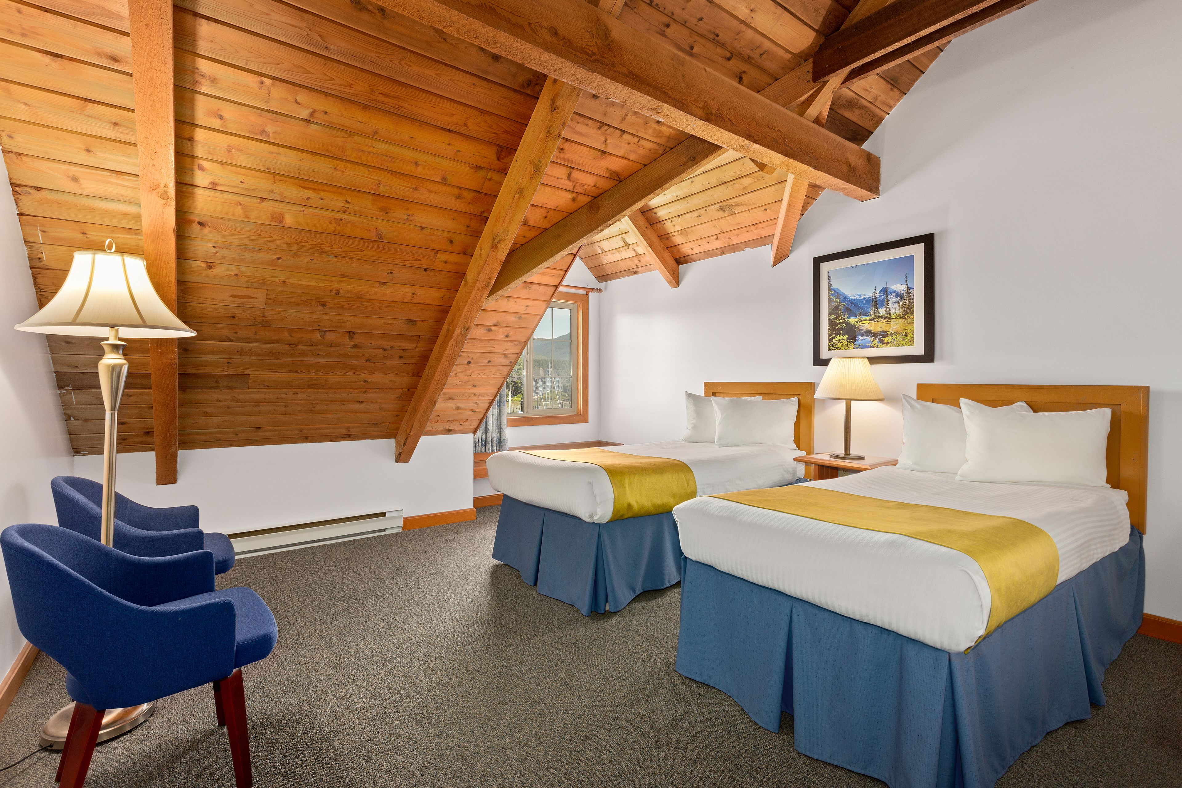 Habitación de hotel con dos camas, una silla azul, vigas de madera en el techo y un cuadro en la pared.