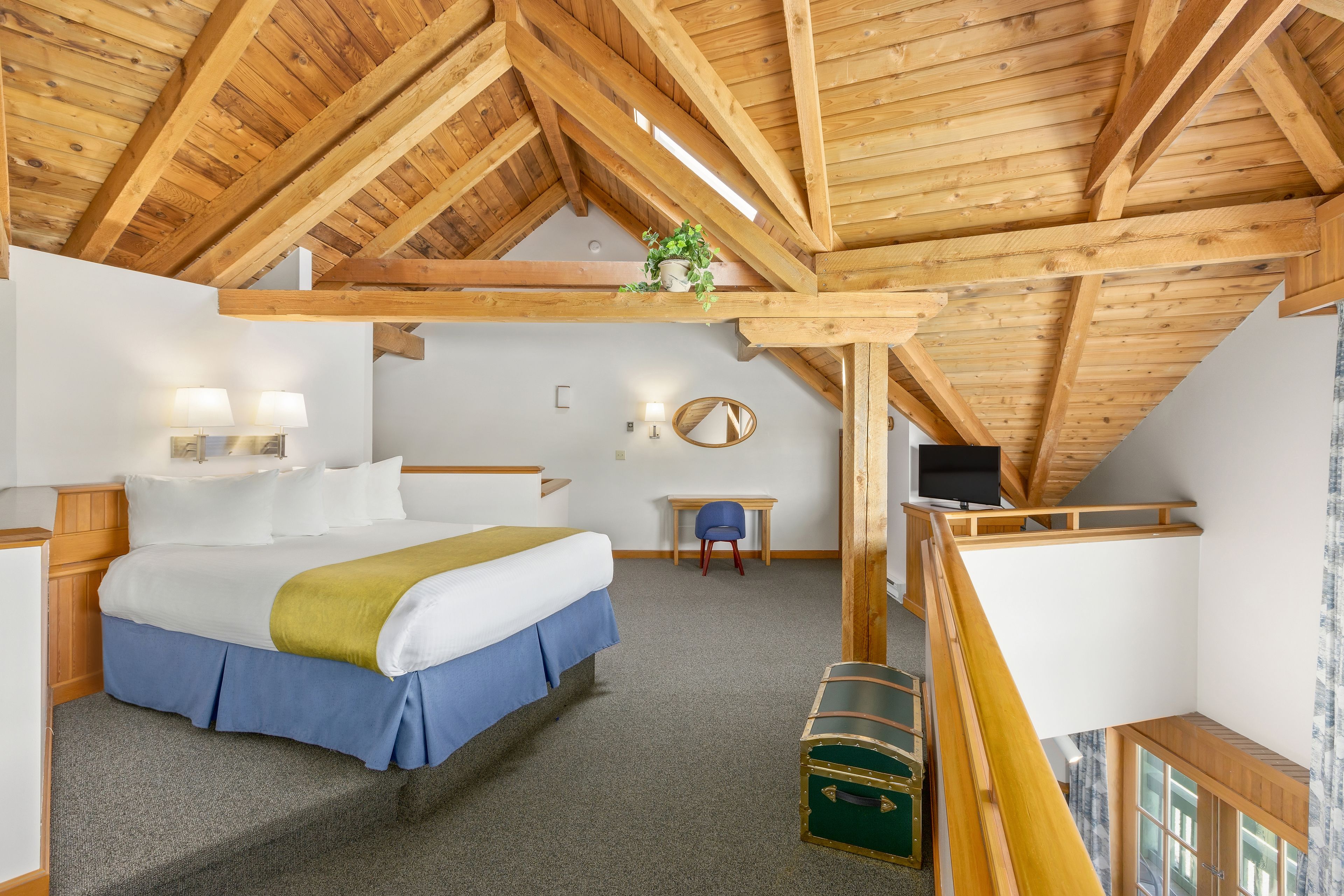 Une chambre avec un plafond en bois, un lit avec une literie blanche et bleue, un bureau avec une chaise et une malle verte.