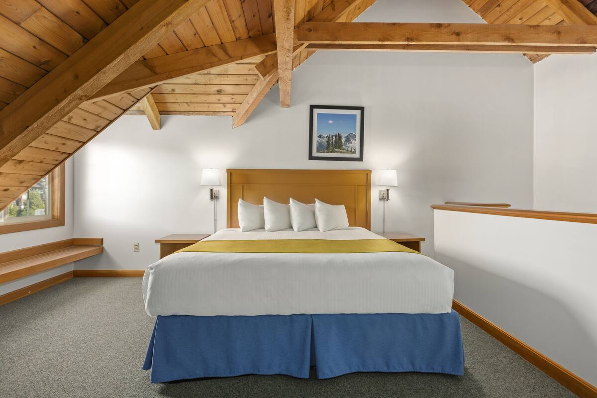 A bedroom with a wooden ceiling, a bed with white and blue bedding, two bedside lamps, and a framed picture on the wall.