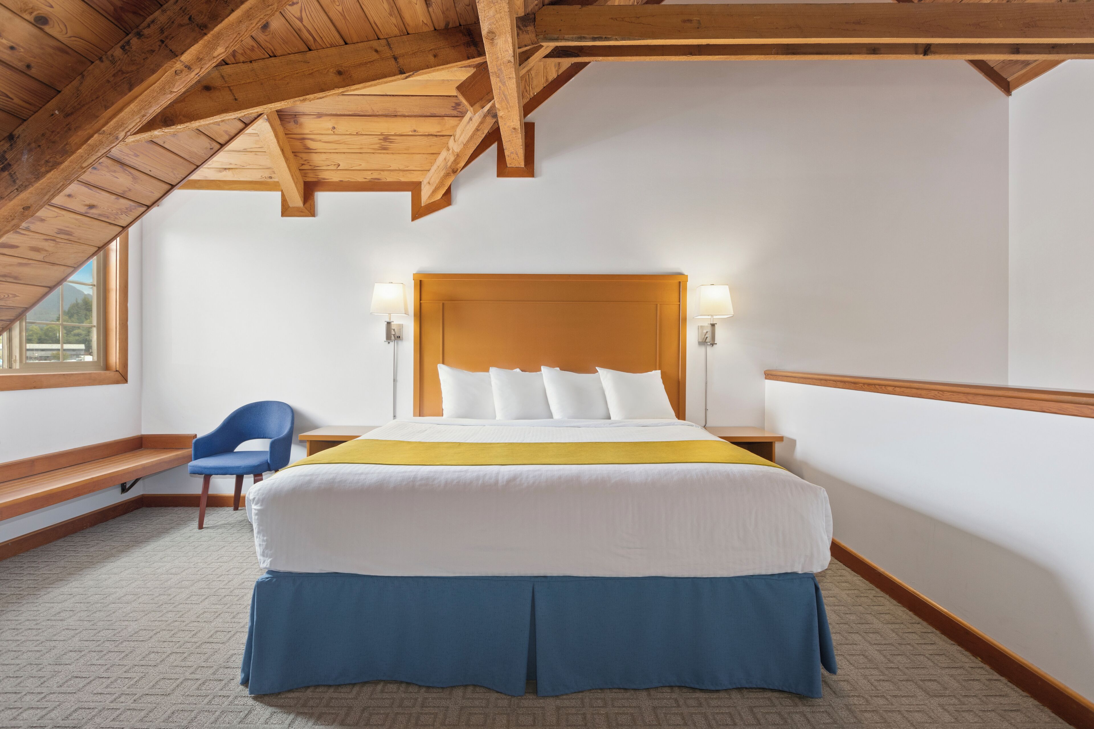 Une chambre d’hôtel avec un grand lit, des poutres en bois au plafond, une chaise bleue et une fenêtre donnant sur un beau paysage.