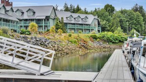A dock with a white railing, a building with multiple windows and a grey roof, and a calm body of water. - Canadian Princess Lodge & Marina (Ucluelet)
