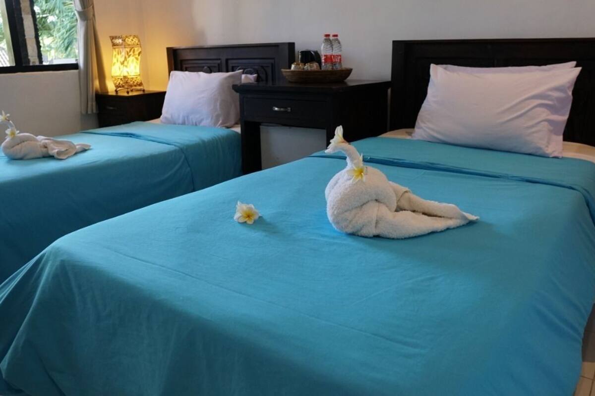 Dos camas individuales con colchas azules, una mesita de noche con lámpara, y una ventana con cortinas.