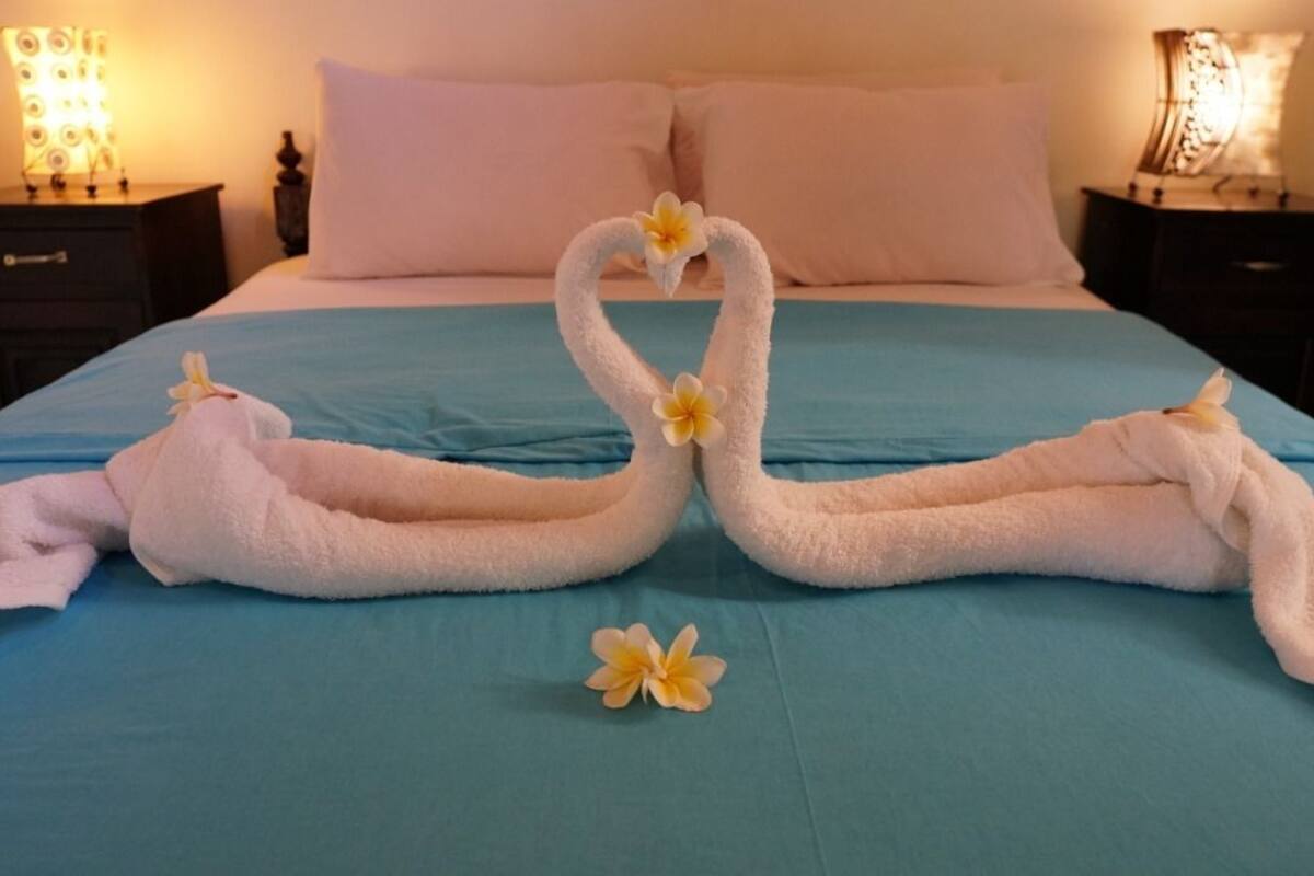 Una cama con cubrecama azul, dos toallas dispuestas en forma de corazón con flores, y dos lámparas de noche.
