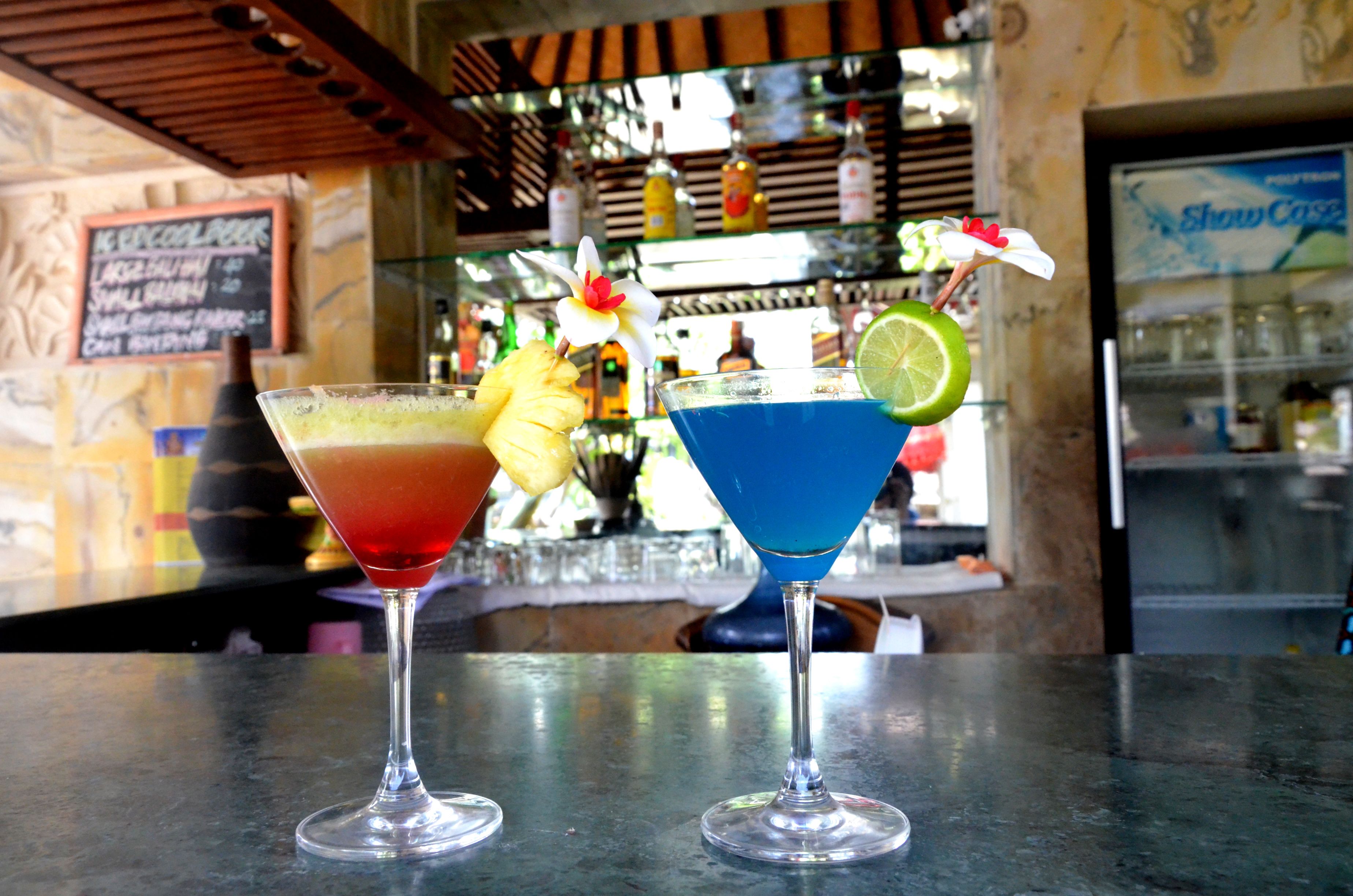 Dois cocktails sobre o balcão de um bar, com um quadro de menu e estantes ao fundo.