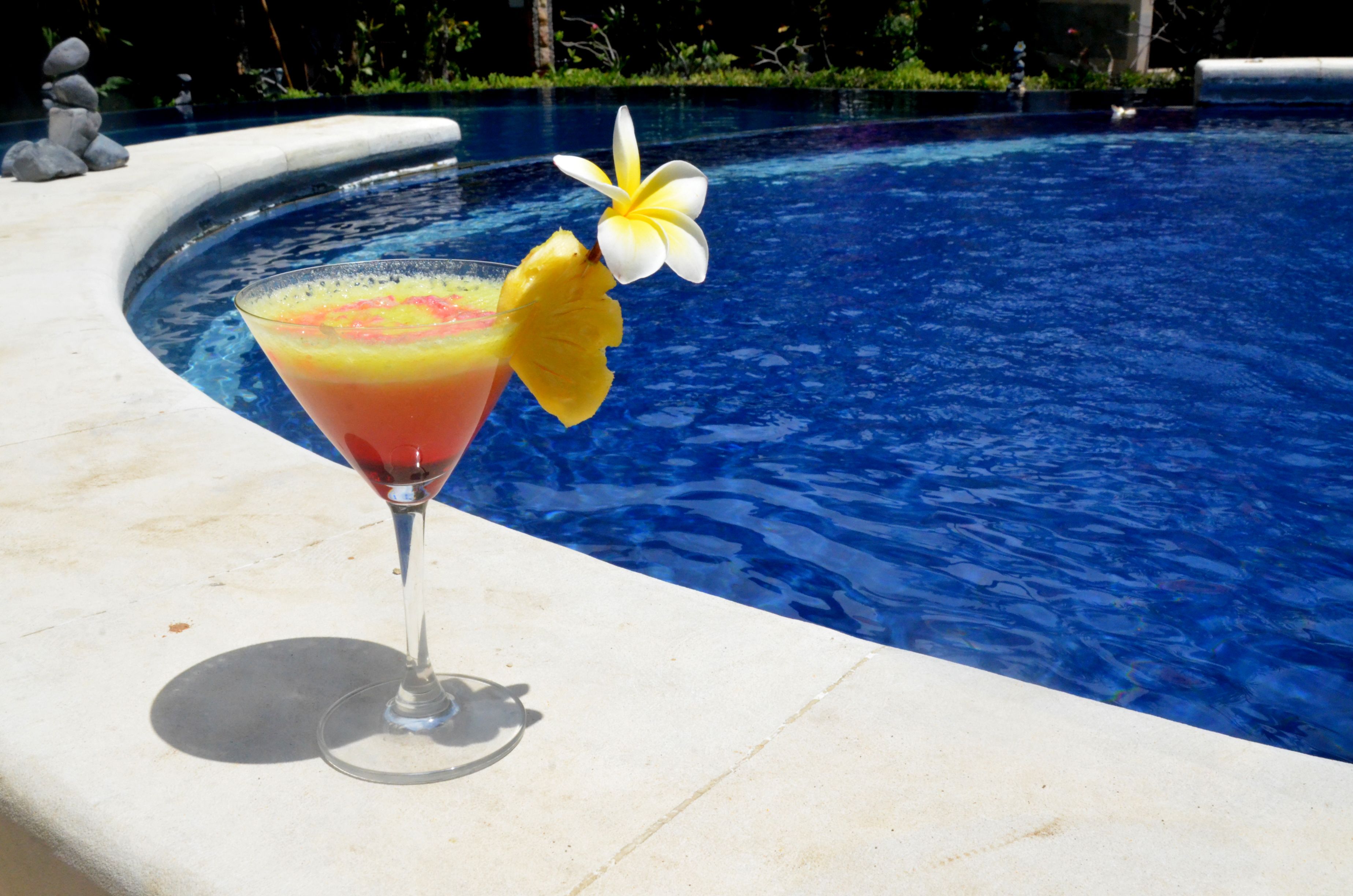 A cocktail in a martini glass with a tropical garnish by a pool.