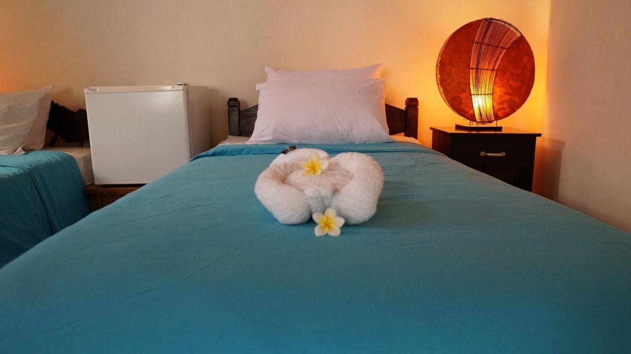Una cama bien tendida con una colcha azul, una almohada blanca y una composición decorativa de toallas con una flor amarilla.