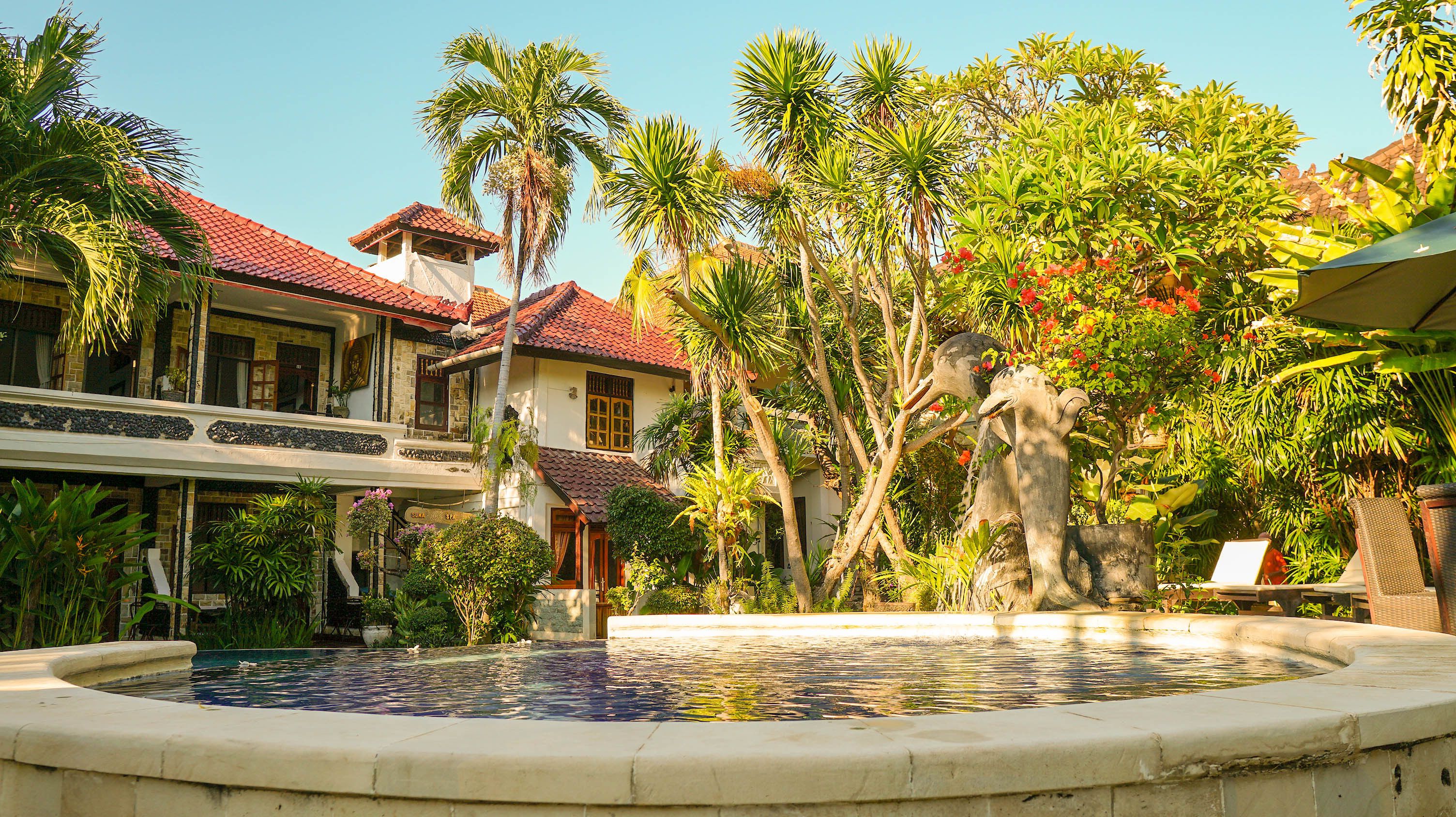 Una piscina con una fuente, rodeada de plantas tropicales y un edificio con techo de tejas rojas.