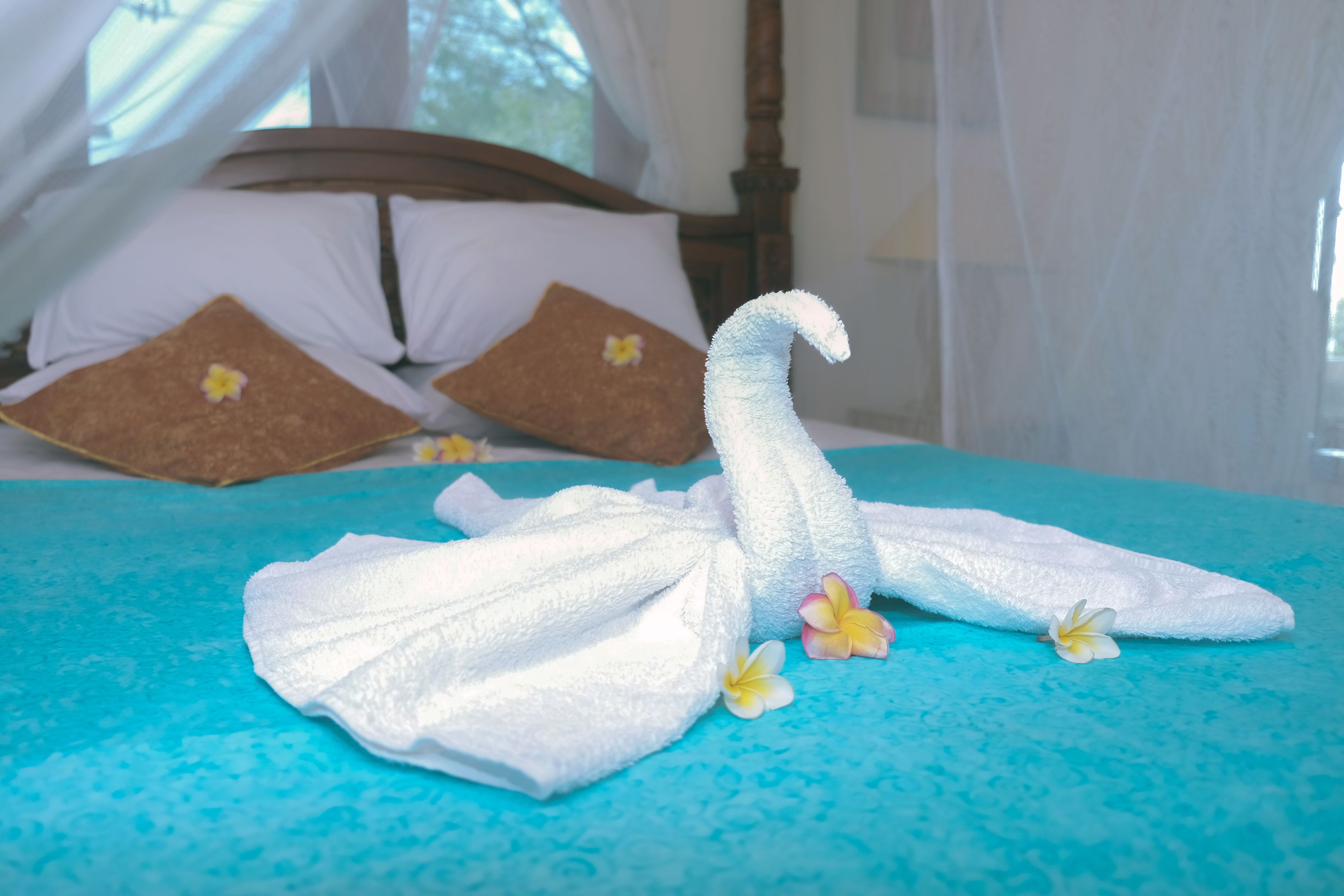 Una cama con cubrecama azul, almohadas blancas y una toalla blanca con forma de cisne decorada con flores.