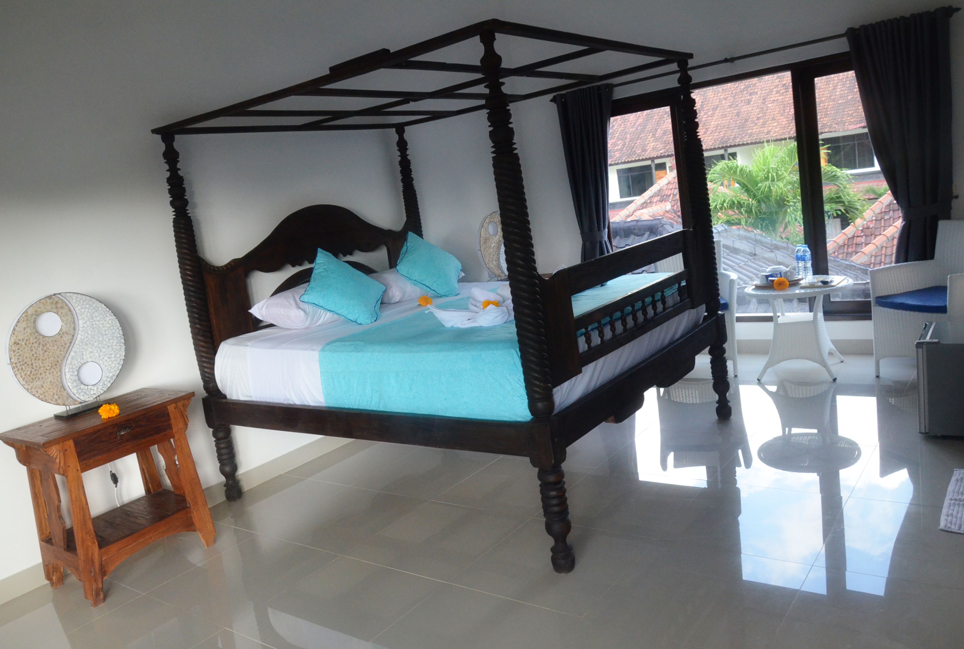 A four-poster bed with a blue bedspread and white linens, a small wooden nightstand with a flower, and a round table with a chair.
