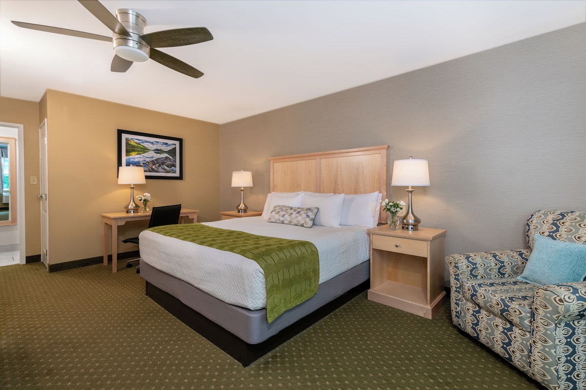 A hotel room with a bed, two bedside tables with lamps, a desk, a chair, and a ceiling fan.