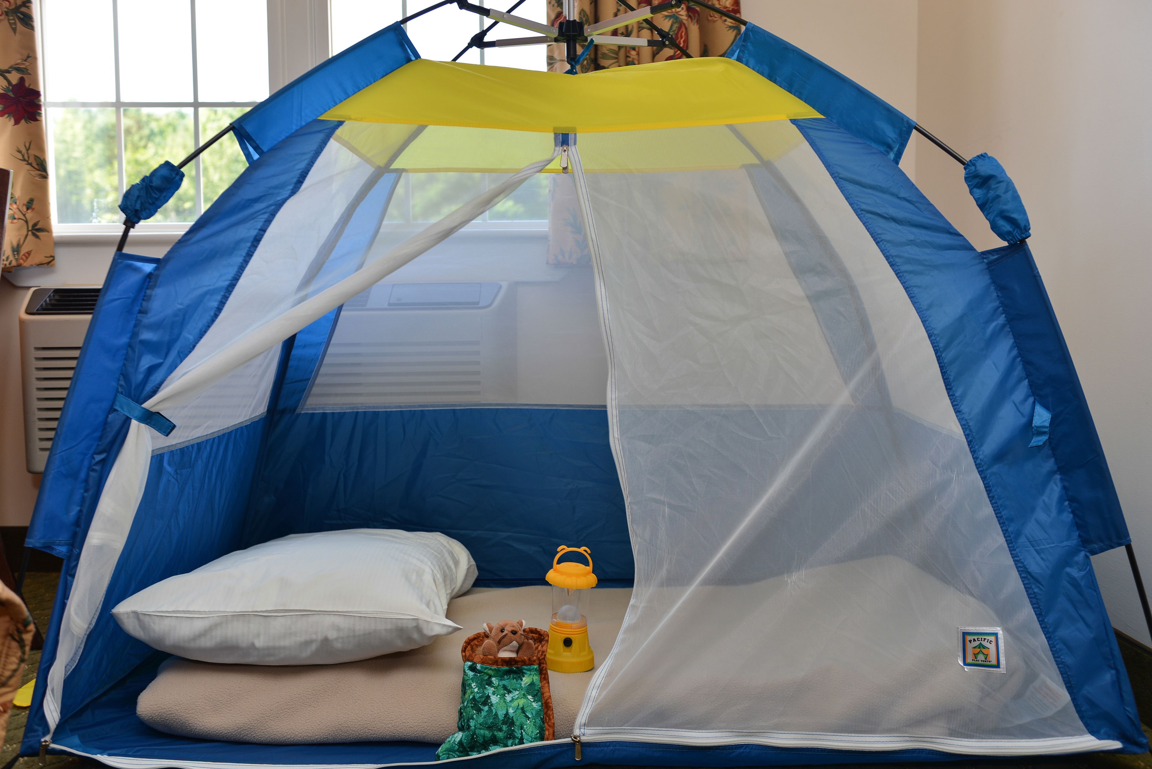 Una carpa azul y blanca con un dosel amarillo, una almohada blanca, una bolsa verde y un farolillo amarillo en su interior.