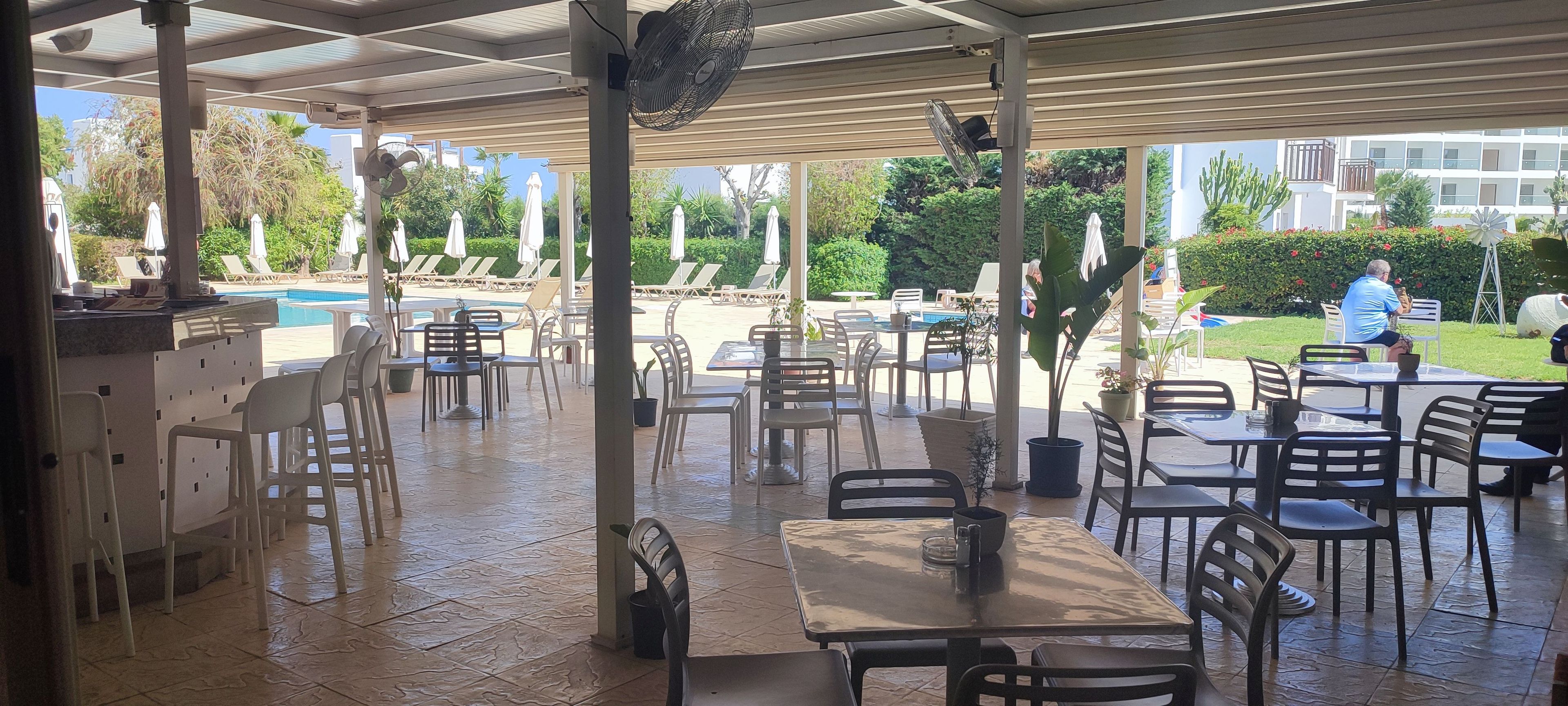 An outdoor patio area with tables and chairs, a pool in the background, and a building with balconies.
