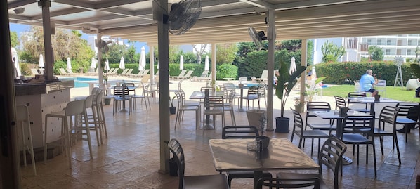 An outdoor patio area with tables and chairs, a pool in the background, and a building with balconies.