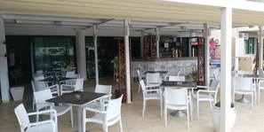 An outdoor cafe area with white plastic chairs and tables, a bar with shelves of bottles, and a view of the city through large windows. - Mandalena Hotel Apts (Paralimni)