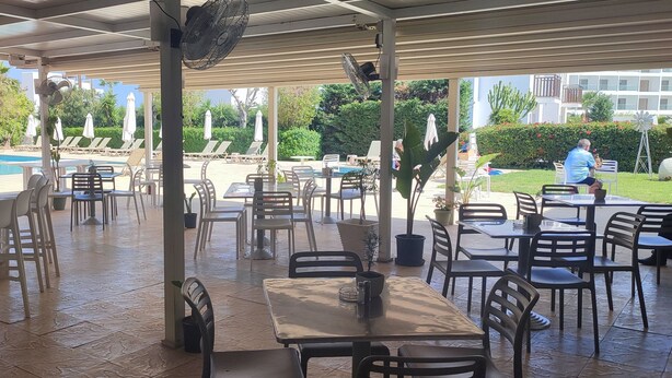 An outdoor patio area with tables and chairs, a pool in the background, and a building with balconies. - Mandalena Hotel Apts (Paralimni)