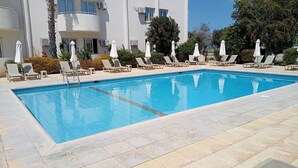 A rectangular swimming pool with tiled edges, surrounded by lounge chairs and umbrellas, set against a building with multiple windows and air conditioning units. - Mandalena Hotel Apts (Paralimni)