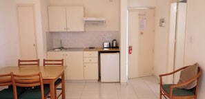 A compact kitchen with a sink, stove, and a small refrigerator. There is a dining table with chairs and a single chair with a cushion. - Mandalena Hotel Apts (Paralimni)