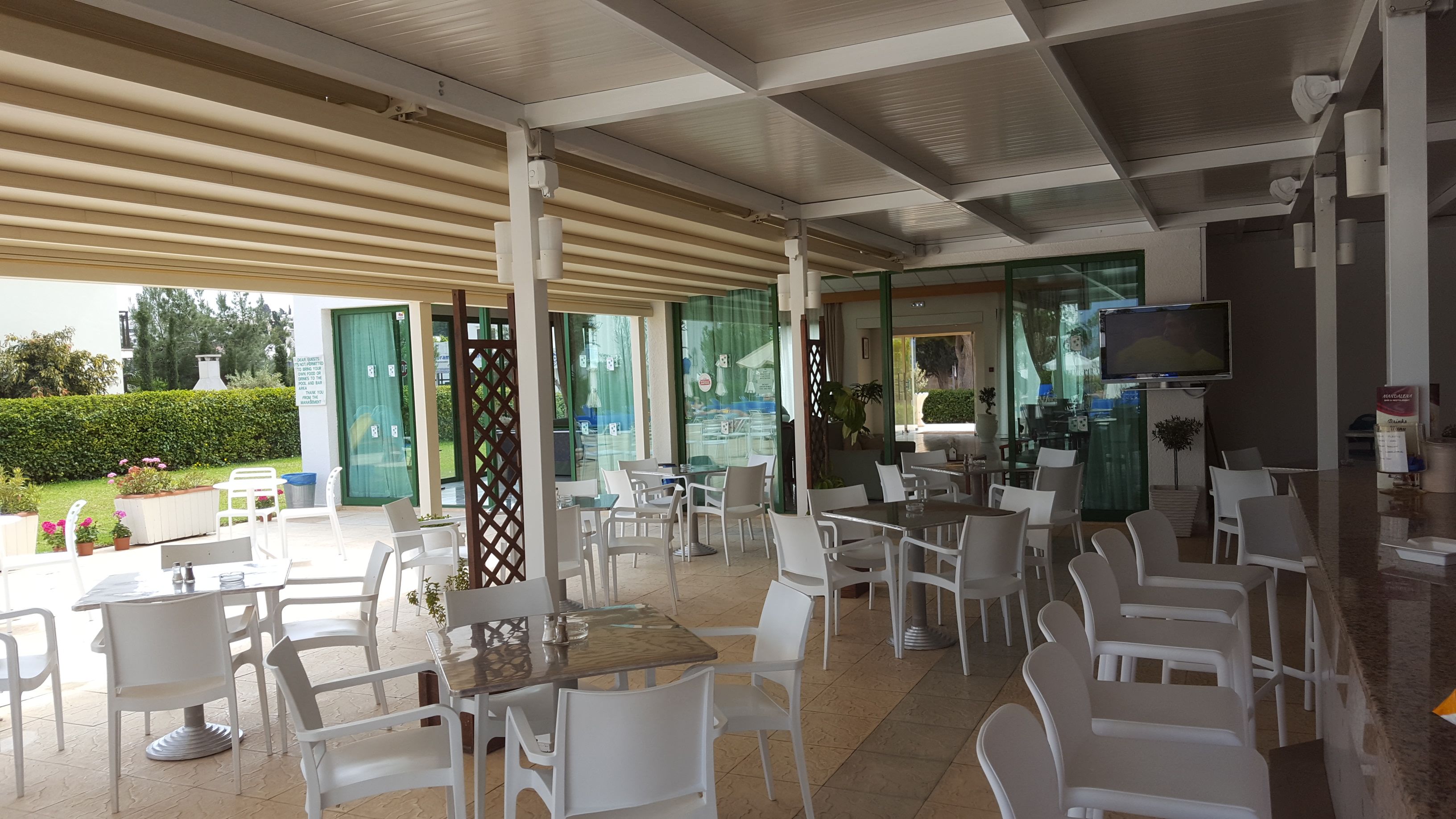 Area caffè all'aperto con sedie e tavoli in plastica bianca, uno schermo televisivo e una parete divisoria in vetro.