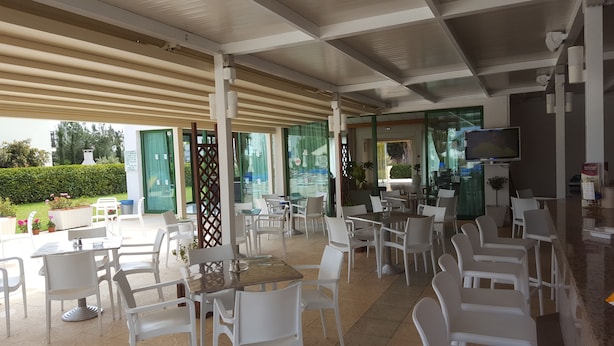 Un área de café al aire libre con sillas y mesas de plástico blanco, una pantalla de televisión y una partición de vidrio.