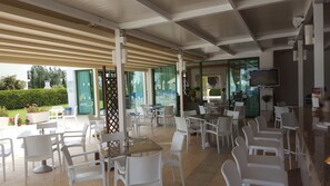An outdoor café area with white plastic chairs and tables, a TV screen, and a glass partition. - Mandalena Hotel Apts (Paralimni)