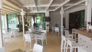 A spacious indoor area with multiple white tables and chairs, a television mounted on the wall, and a reception desk with a menu. - Mandalena Hotel Apts (Paralimni)
