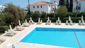 Piscina rectangular con bordes de azulejos, rodeada de sillas de descanso y sombrillas, con edificios residenciales y vegetación de fondo.