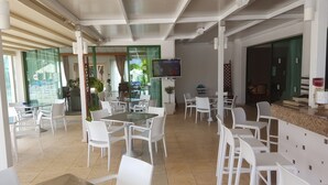 A spacious indoor area with multiple white tables and chairs, a television mounted on the wall, and a reception desk with a menu. - Mandalena Hotel Apts (Paralimni)