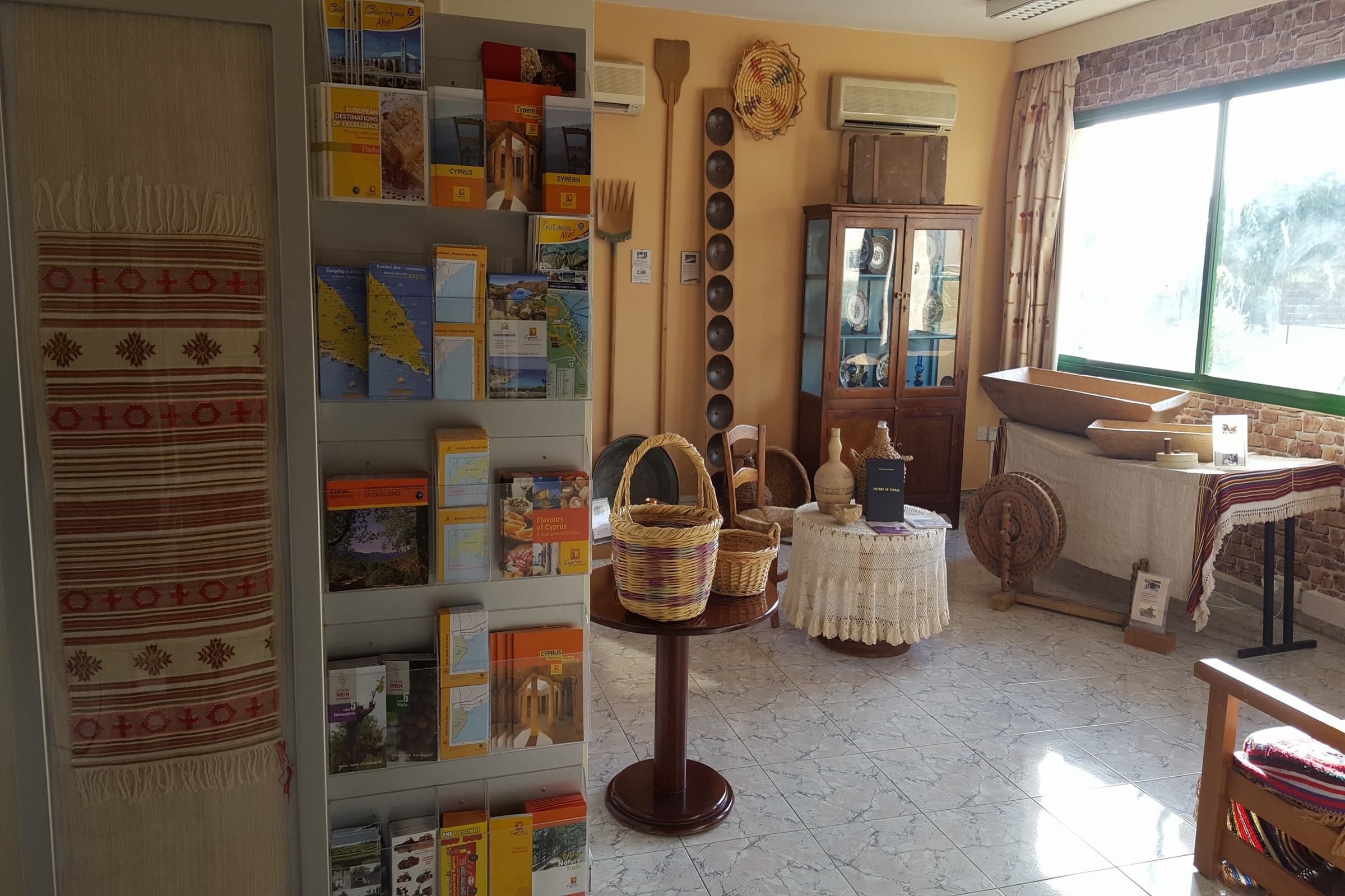 A room with a bookshelf, wicker baskets, a spinning wheel, and a window with curtains.