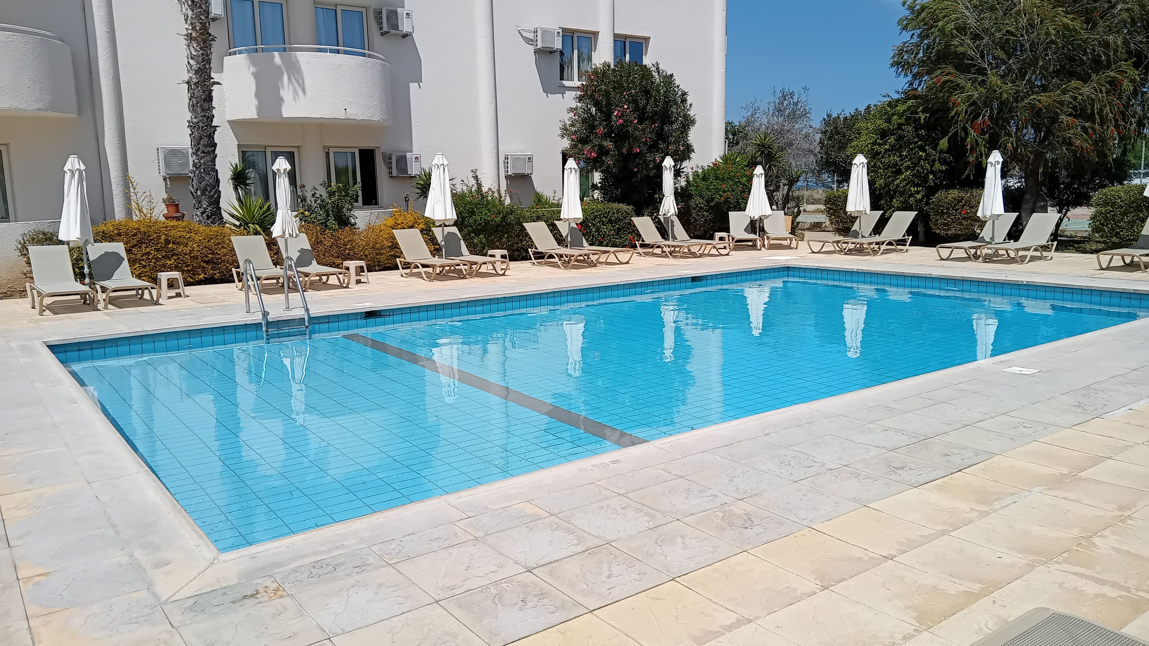 A rectangular swimming pool with tiled edges, surrounded by lounge chairs and umbrellas, set against a building with multiple windows and air conditioning units.