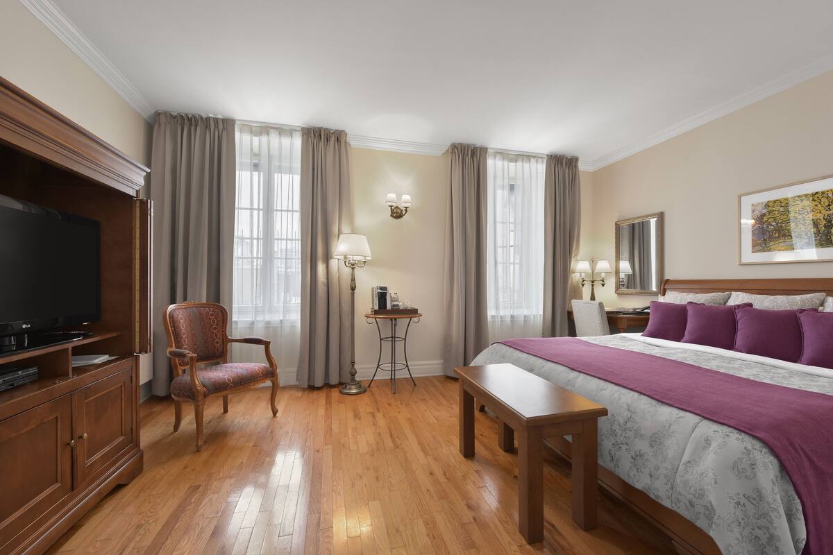 A hotel room with a wooden floor, a bed with a purple blanket, a TV on a wooden cabinet, a chair, a small table with a lamp, and a painting on the wall.