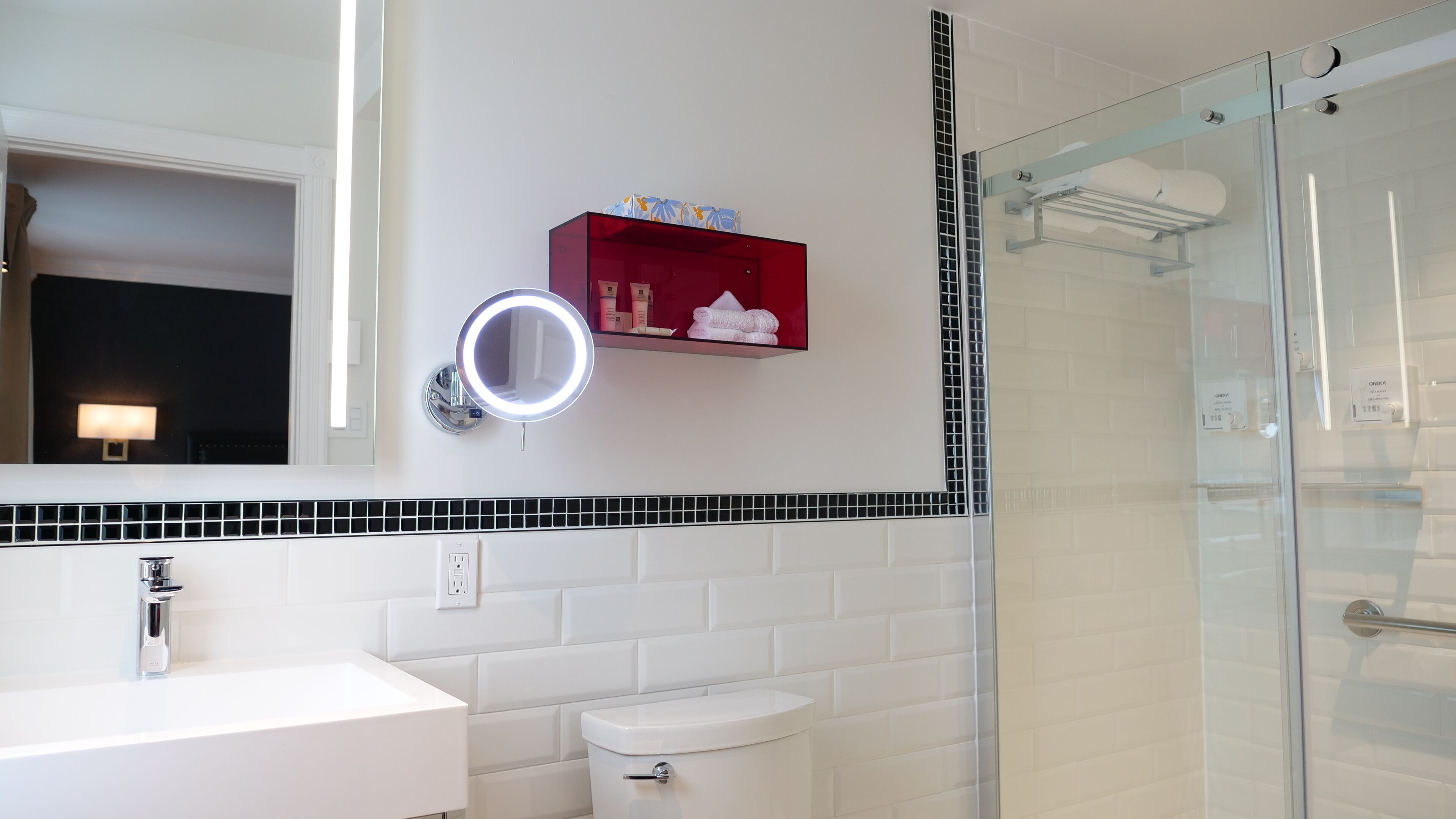 Un bany modern amb un lavabo blanc, un box de dutxa de vidre, un suport de tovalloles vermell i un mirall amb llum.