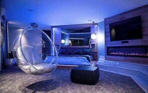 A modern bedroom with a hanging chair, a bed, a TV, and a fireplace. - Le Fabreville (Laval)